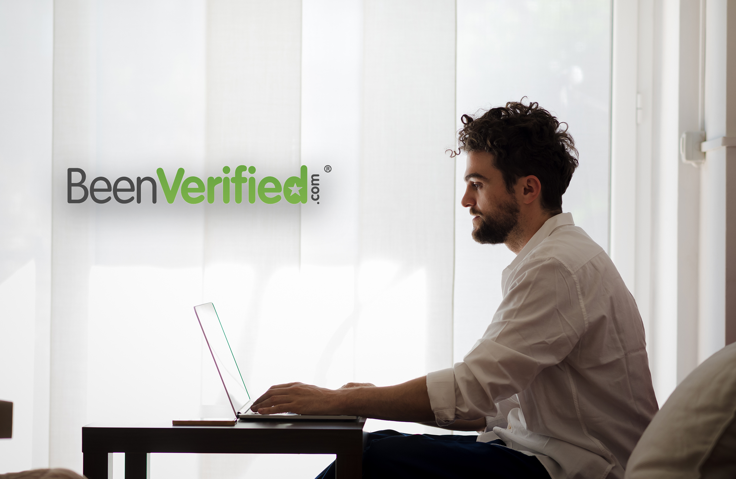 A man with curly hair sits at a desk using a laptop in a bright office with the BeenVerified logo on the wall behind him.